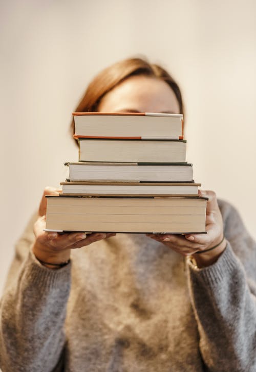 Anonymous woman in soft sweater covering face with pile of books on light background