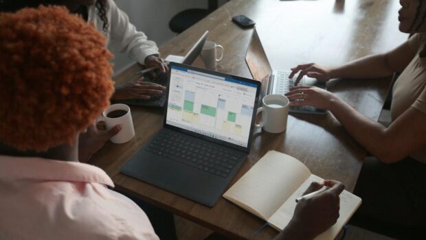 Three people in a meeting at a table discussing schedule on their Microsoft laptop